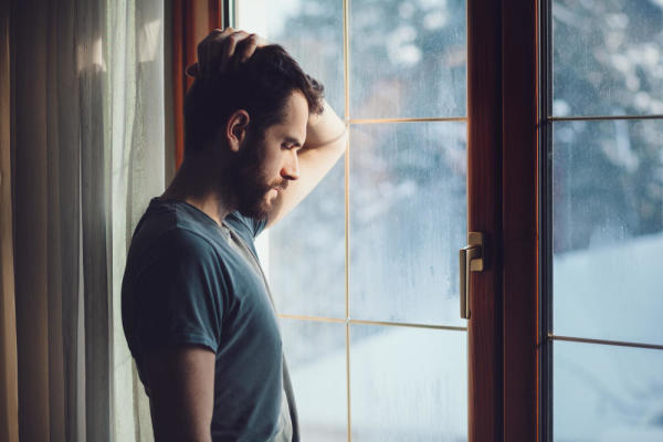 Man stands by a window with his hand on his head, frustrated by his substance use disorder